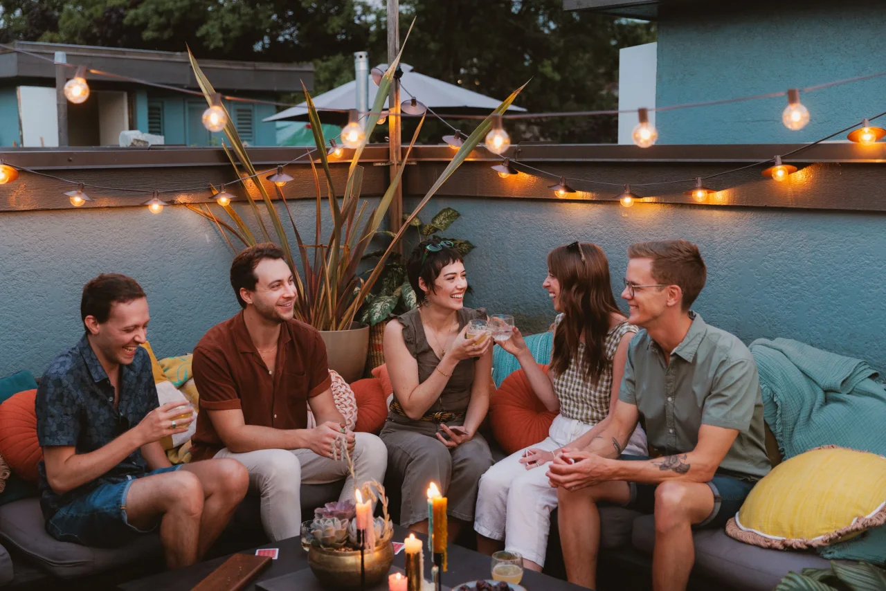 Cozy backyard patio at night strung with warm glow string lights, where friends lounge on pillows and blankets watching a movie on a projector screen, surrounded by plants, candles, and rattan chairs.